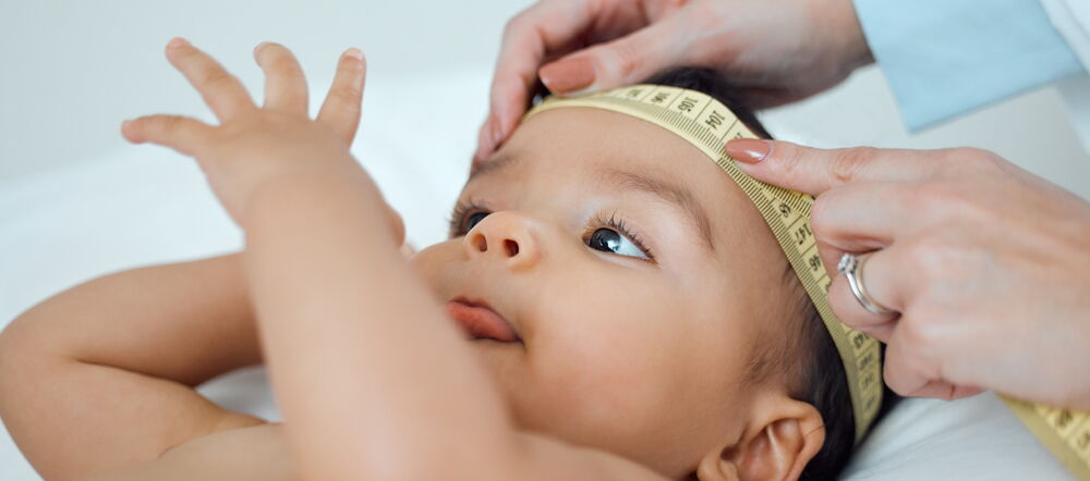 A baby lies on a white surface while a hand with a measuring tape measures the baby's head.