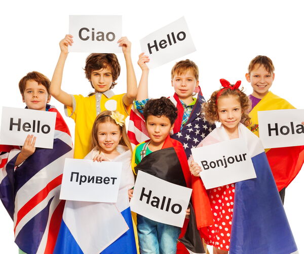 Nine children hold posters with greetings in different languages: ‘Ciao’, ‘Hello’, ‘Hola’, ‘Bonjour’, ‘Hallo’, ‘Привет’. They wear colorful clothes and capes with flags.