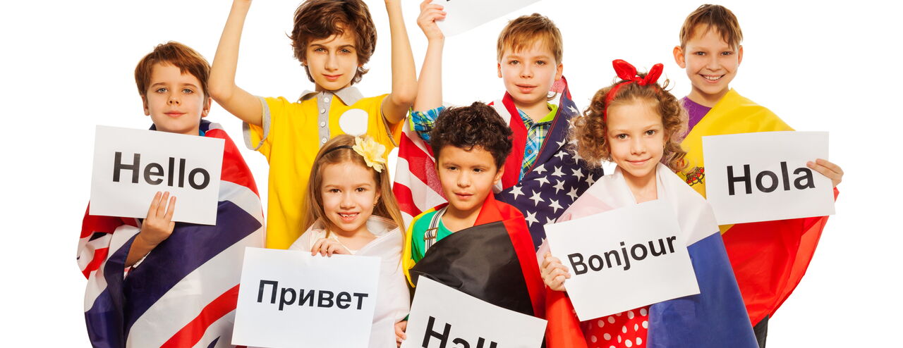 Nine children hold posters with greetings in different languages: ‘Ciao’, ‘Hello’, ‘Hola’, ‘Bonjour’, ‘Hallo’, ‘Привет’. They wear colorful clothes and capes with flags.