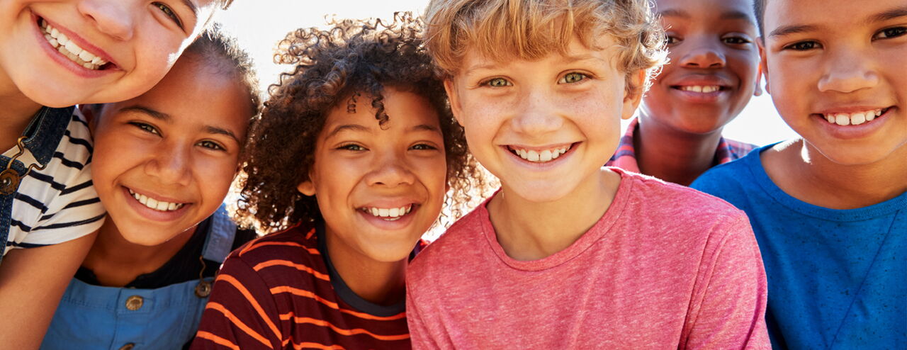 Six children with different hairstyles and skin tones standing together in casual clothes. They are smiling and posing for a photo.