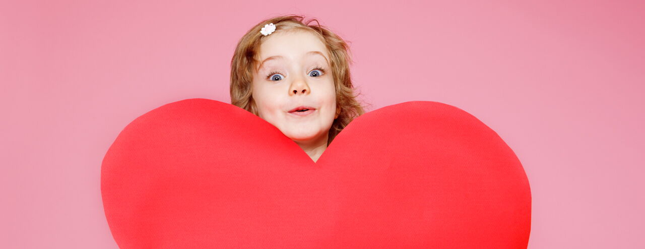 A girl is holding a large red fabric heart in front of her. The background is pink.